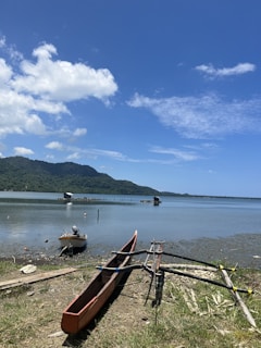 A tranquil lakeside scene with a traditional canoe and small boat resting on the shore. The water is calm with distant fish huts extending into the lake. Lush green mountains form the backdrop under a clear blue sky with scattered clouds.
