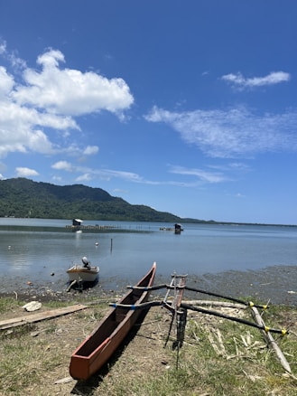 A tranquil lakeside scene with a traditional canoe and small boat resting on the shore. The water is calm with distant fish huts extending into the lake. Lush green mountains form the backdrop under a clear blue sky with scattered clouds.