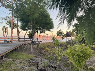 A wooden boardwalk leads through a natural setting, bordered by lush green trees and plants. A large butterfly-shaped structure is visible to the left. In the background, there is a sign displaying 'Pesona Indonesia', framed by trees. The setting appears to be near water, with the horizon visible in the distance.
