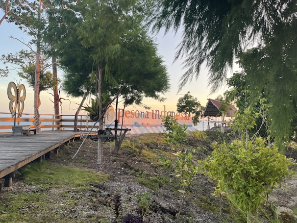 A wooden boardwalk leads through a natural setting, bordered by lush green trees and plants. A large butterfly-shaped structure is visible to the left. In the background, there is a sign displaying 'Pesona Indonesia', framed by trees. The setting appears to be near water, with the horizon visible in the distance.