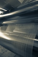 Large industrial metal cylinders and tightly wound wire or thread are arranged in parallel. Strong linear patterns and reflections are visible, suggesting a machine or manufacturing setting.