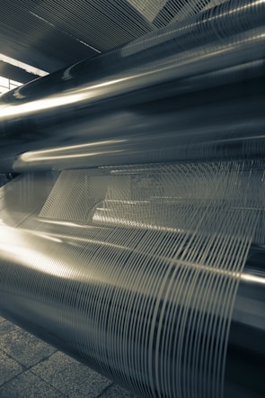 Large industrial metal cylinders and tightly wound wire or thread are arranged in parallel. Strong linear patterns and reflections are visible, suggesting a machine or manufacturing setting.