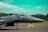 A military jet fighter is stationed on an airstrip with a clear blue sky and lush greenery in the background. The aircraft is sleek and gray, with visible details on the fuselage and cockpit. The runway surface is marked with yellow lines, and there are sandbags placed near the aircraft.