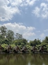 A row of small wooden cabins sits along the edge of a calm lake, surrounded by lush green trees and foliage. The sky above is dotted with fluffy white clouds, suggesting a clear, peaceful day.