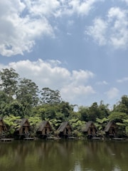 A row of small wooden cabins sits along the edge of a calm lake, surrounded by lush green trees and foliage. The sky above is dotted with fluffy white clouds, suggesting a clear, peaceful day.