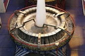 A large circular industrial weaving machine with numerous threads being woven into a fabric. The machine has a metallic structure with clamps and handles, and it is placed on a checkered metal floor. The threads are in motion, suggesting that the machine is actively weaving.