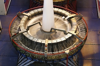 A large circular industrial weaving machine with numerous threads being woven into a fabric. The machine has a metallic structure with clamps and handles, and it is placed on a checkered metal floor. The threads are in motion, suggesting that the machine is actively weaving.