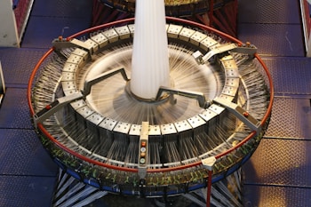 A large circular industrial weaving machine with numerous threads being woven into a fabric. The machine has a metallic structure with clamps and handles, and it is placed on a checkered metal floor. The threads are in motion, suggesting that the machine is actively weaving.