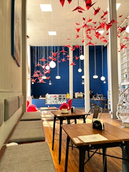 A cozy cafe interior with modern decor, featuring wooden tables and benches with gray cushions. Red origami cranes hang from the ceiling, complementing the blue walls that have a textured design. There are circular pendant lights hanging amidst the origami cranes. The cafe has a small counter and display case, with red chairs adding a vibrant touch.