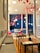 A cozy cafe interior with modern decor, featuring wooden tables and benches with gray cushions. Red origami cranes hang from the ceiling, complementing the blue walls that have a textured design. There are circular pendant lights hanging amidst the origami cranes. The cafe has a small counter and display case, with red chairs adding a vibrant touch.