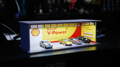 A miniature display resembling a Shell V-Power gas station is shown, featuring four model cars parked in front. The backdrop includes a Shell logo and the words 'Shell V-Power' in red with a yellow and red color scheme.
