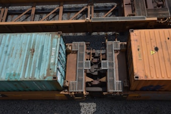 A top-down view of two shipping containers on a train, with a rusty orange color and visible structural metal parts connecting them. One container is blue and the other is orange; they are positioned on gravel tracks.