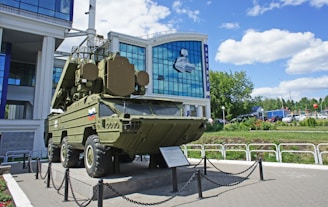 A military vehicle is displayed outside a modern building with large glass windows. The vehicle is olive green and mounted on a platform surrounded by a chain barrier. There are plaques or informational signs placed in front of it. The building behind has a mural depicting a spacecraft and the word 'Vostok' written in Cyrillic. The sky is partly cloudy, and there are some cars and greenery in the background.
