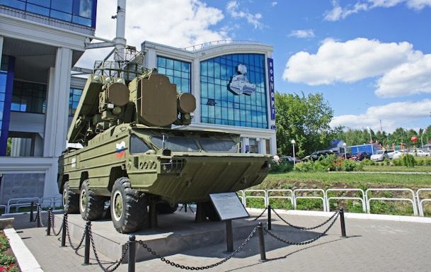 A military vehicle is displayed outside a modern building with large glass windows. The vehicle is olive green and mounted on a platform surrounded by a chain barrier. There are plaques or informational signs placed in front of it. The building behind has a mural depicting a spacecraft and the word 'Vostok' written in Cyrillic. The sky is partly cloudy, and there are some cars and greenery in the background.