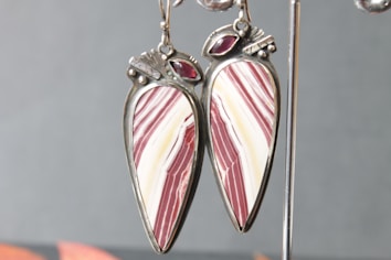 A pair of dangling earrings with a teardrop shape, featuring a polished design of red and white stripes. The earrings are set in metallic frames and adorned with small, red gemstone accents at the top.