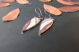 A pair of teardrop-shaped earrings with maroon and white stripes is placed on a dark textured surface surrounded by scattered autumn leaves in shades of orange and red. The earrings have metal hooks and an elegant, polished look.