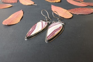 A pair of teardrop-shaped earrings with maroon and white stripes is placed on a dark textured surface surrounded by scattered autumn leaves in shades of orange and red. The earrings have metal hooks and an elegant, polished look.
