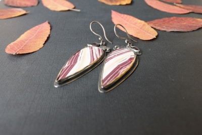 A pair of teardrop-shaped earrings with maroon and white stripes is placed on a dark textured surface surrounded by scattered autumn leaves in shades of orange and red. The earrings have metal hooks and an elegant, polished look.