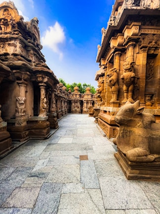 An intricately carved stone temple features ornate pillars and sculptures, creating a pathway through the historical architecture. The temple surfaces are adorned with detailed carvings of deities and mythical creatures, set against a clear blue sky and lush green foliage in the background.