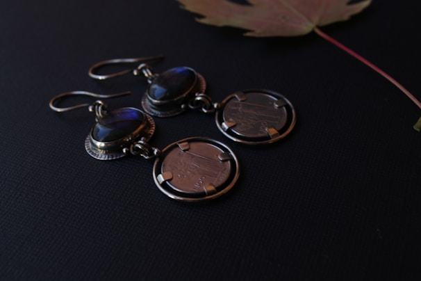 A pair of earrings featuring oval gemstones with a deep blue hue are attached to circular metal discs resembling coins. The earrings are placed on a dark surface next to a dried maple leaf, which adds a subtle natural element to the composition.