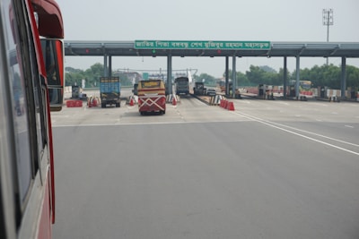 A highway toll plaza featuring several lanes for vehicles to pass through. Red and white traffic cones and barriers are placed around the lanes, and vehicles, including buses and trucks, are waiting to move forward. There is a sign with text on the structure overhead.