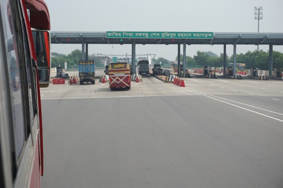 A highway toll plaza featuring several lanes for vehicles to pass through. Red and white traffic cones and barriers are placed around the lanes, and vehicles, including buses and trucks, are waiting to move forward. There is a sign with text on the structure overhead.