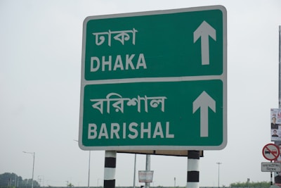 A roadside sign with directions to Dhaka and Barishal in both English and Bengali languages. The sign is green with white text and arrows pointing upwards. It is mounted on a pole with black and white stripes. In the background, there are faint outlines of other signposts and a small piece of a poster on the right.