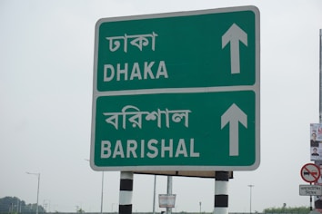 A roadside sign with directions to Dhaka and Barishal in both English and Bengali languages. The sign is green with white text and arrows pointing upwards. It is mounted on a pole with black and white stripes. In the background, there are faint outlines of other signposts and a small piece of a poster on the right.