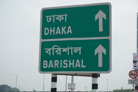 A roadside sign with directions to Dhaka and Barishal in both English and Bengali languages. The sign is green with white text and arrows pointing upwards. It is mounted on a pole with black and white stripes. In the background, there are faint outlines of other signposts and a small piece of a poster on the right.