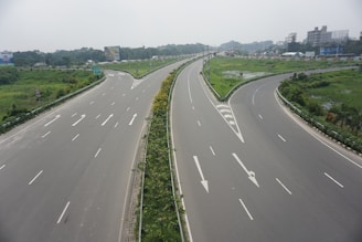 A wide highway intersection with multiple lanes and clear white road markings. The roads are bordered by lush green grass and some trees, indicating a well-maintained area. Traffic signs and billboards are visible in the background, suggesting an urban or semi-urban setting. The sky is overcast, giving a muted lighting to the scene.
