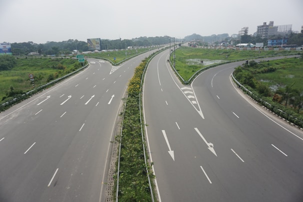 A wide highway intersection with multiple lanes and clear white road markings. The roads are bordered by lush green grass and some trees, indicating a well-maintained area. Traffic signs and billboards are visible in the background, suggesting an urban or semi-urban setting. The sky is overcast, giving a muted lighting to the scene.
