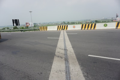 An empty highway intersection with a clear view of the road and surrounding landscape. The pavement shows a visible seam running down the middle. A barrier with distinctive black and yellow stripes is present, adorned with several circular emblems. In the distance, industrial buildings and advertising billboards are visible, along with a tall streetlight.