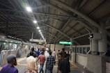 A busy subway or train station platform with commuters walking in different directions. The platform is under a large arched roof structure with metal beams. There is a train on the left side with its doors open, and signs with directions and train information are hanging from the ceiling.