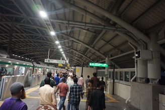 A busy subway or train station platform with commuters walking in different directions. The platform is under a large arched roof structure with metal beams. There is a train on the left side with its doors open, and signs with directions and train information are hanging from the ceiling.