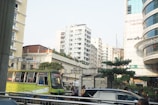 Urban scene featuring several multi-story buildings with a mix of concrete and glass. A worn-out city bus with bright yellow and green paint is in the foreground, surrounded by a street bustling with people and cars. The sky is clear and adds a bright ambiance to the scene.