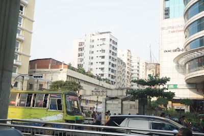 Urban scene featuring several multi-story buildings with a mix of concrete and glass. A worn-out city bus with bright yellow and green paint is in the foreground, surrounded by a street bustling with people and cars. The sky is clear and adds a bright ambiance to the scene.
