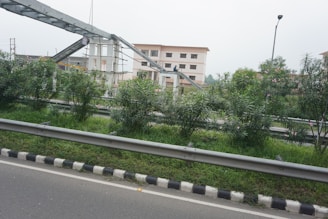The image features an urban setting with a road bordered by metal railings and a neatly maintained green median with bushes. In the background, there is an under-construction pedestrian overpass connected to two multi-story buildings. A single streetlight is also visible.