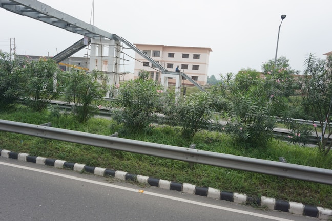 The image features an urban setting with a road bordered by metal railings and a neatly maintained green median with bushes. In the background, there is an under-construction pedestrian overpass connected to two multi-story buildings. A single streetlight is also visible.
