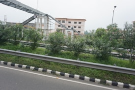 The image features an urban setting with a road bordered by metal railings and a neatly maintained green median with bushes. In the background, there is an under-construction pedestrian overpass connected to two multi-story buildings. A single streetlight is also visible.