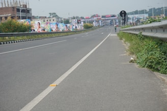 A wide highway with minimal traffic, featuring road signs and a pedestrian walking along the side. Billboards with colorful images line the road, and greenery grows beside the guardrails.