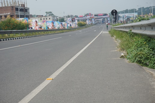 A wide highway with minimal traffic, featuring road signs and a pedestrian walking along the side. Billboards with colorful images line the road, and greenery grows beside the guardrails.