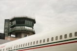 A control tower stands prominently with multiple windows and antennae on top. The adjacent terminal building and part of an airplane's fuselage are visible, characterized by a white exterior with green and red stripes.