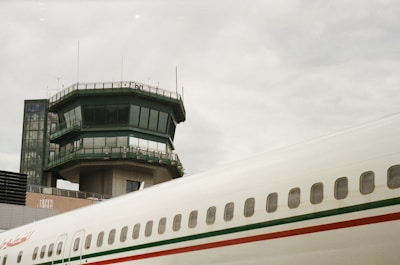 A control tower stands prominently with multiple windows and antennae on top. The adjacent terminal building and part of an airplane's fuselage are visible, characterized by a white exterior with green and red stripes.