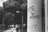 A black and white photograph captures a cityscape scene. In the foreground, graffiti artwork featuring a simple, cartoonish figure with the word 'A?TIST' is prominently drawn on a large cylindrical pillar. Behind the pillar, two people are walking down a tree-lined street, giving a sense of urban life. The scene has a slightly overcast, moody atmosphere with street lamps arching over the path.