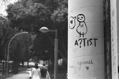 A black and white photograph captures a cityscape scene. In the foreground, graffiti artwork featuring a simple, cartoonish figure with the word 'A?TIST' is prominently drawn on a large cylindrical pillar. Behind the pillar, two people are walking down a tree-lined street, giving a sense of urban life. The scene has a slightly overcast, moody atmosphere with street lamps arching over the path.