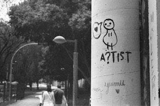 A black and white photograph captures a cityscape scene. In the foreground, graffiti artwork featuring a simple, cartoonish figure with the word 'A?TIST' is prominently drawn on a large cylindrical pillar. Behind the pillar, two people are walking down a tree-lined street, giving a sense of urban life. The scene has a slightly overcast, moody atmosphere with street lamps arching over the path.