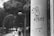 A black and white photograph captures a cityscape scene. In the foreground, graffiti artwork featuring a simple, cartoonish figure with the word 'A?TIST' is prominently drawn on a large cylindrical pillar. Behind the pillar, two people are walking down a tree-lined street, giving a sense of urban life. The scene has a slightly overcast, moody atmosphere with street lamps arching over the path.