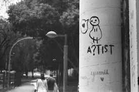 A black and white photograph captures a cityscape scene. In the foreground, graffiti artwork featuring a simple, cartoonish figure with the word 'A?TIST' is prominently drawn on a large cylindrical pillar. Behind the pillar, two people are walking down a tree-lined street, giving a sense of urban life. The scene has a slightly overcast, moody atmosphere with street lamps arching over the path.