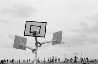 A black and white photograph featuring a unique basketball hoop structure with three backboards in an open area, surrounded by people. In the background, a group of individuals are gathered near a volleyball net, with more people dispersed, engaging in recreational activities.