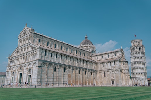 A grand architectural complex featuring an ornate cathedral with decorative arches and columns, accompanied by a recognizable leaning bell tower. A clear blue sky and green grass enhance the scene, with tourists walking around the area.
